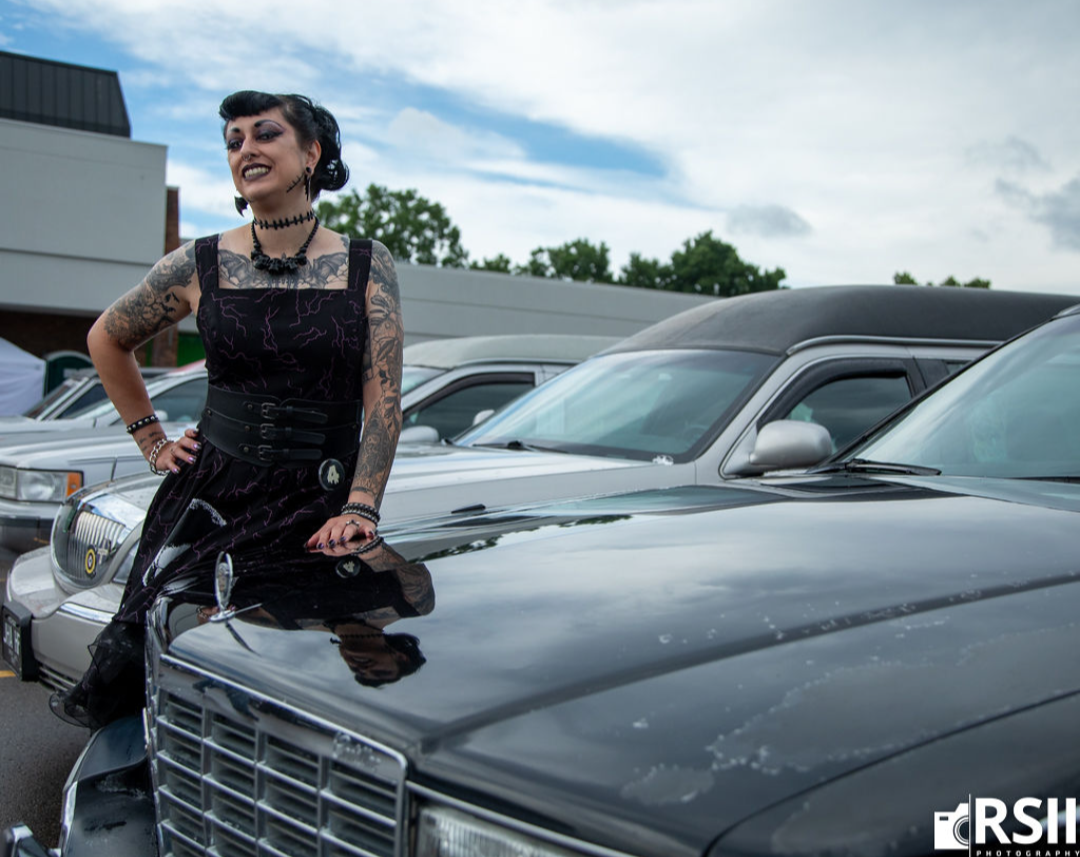 A woman with dark hair and tattoos leans against the hood of a black car in an outdoor parking lot. She is wearing a black lace-trimmed corset-style dress with a matching corset-style waist belt and black boots.