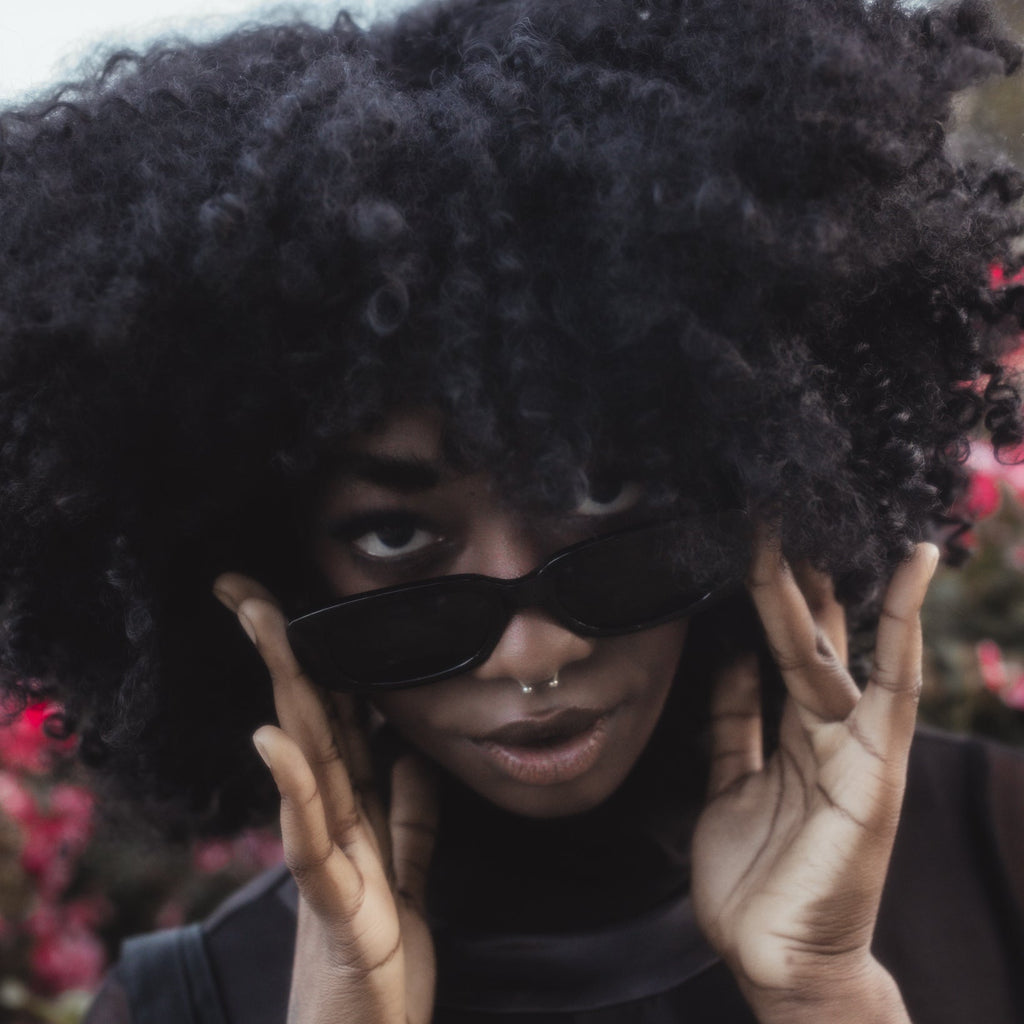 Person with an afro wearing sunglasses and holding hands to face, with blurred background