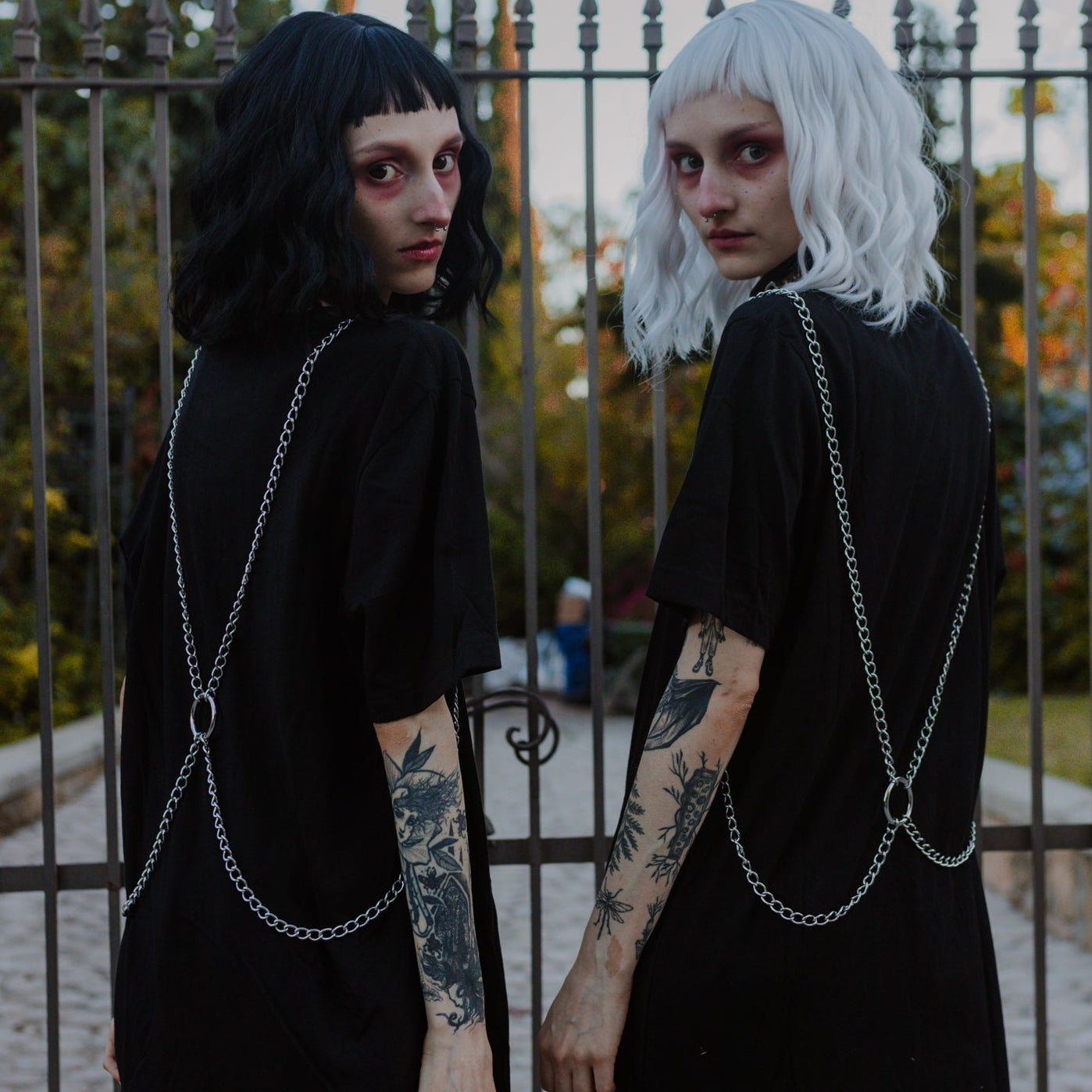 Two individuals wearing black dresses with chain details in front of a metal fence.