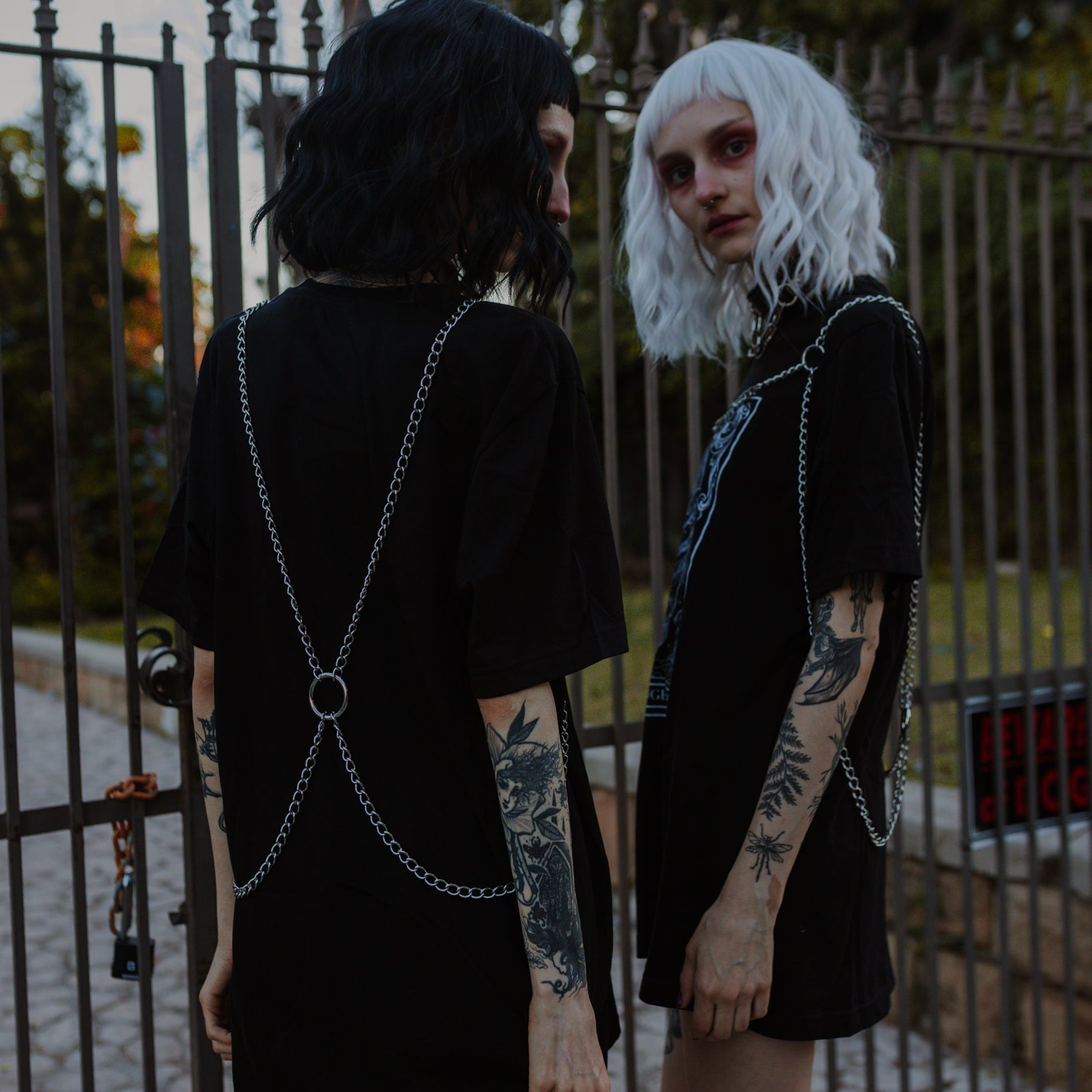 Two individuals in black outfits with chain details standing in front of a metal gate.