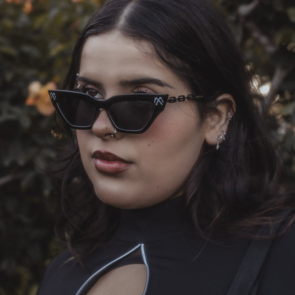 Woman wearing black sunglasses and a black top with a cut-out design, standing against a blurred natural background.