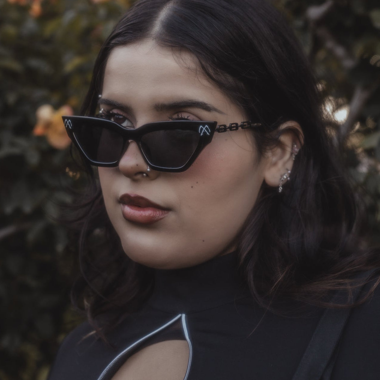 Woman wearing black sunglasses and a black top with a cut-out design, standing against a blurred natural background.