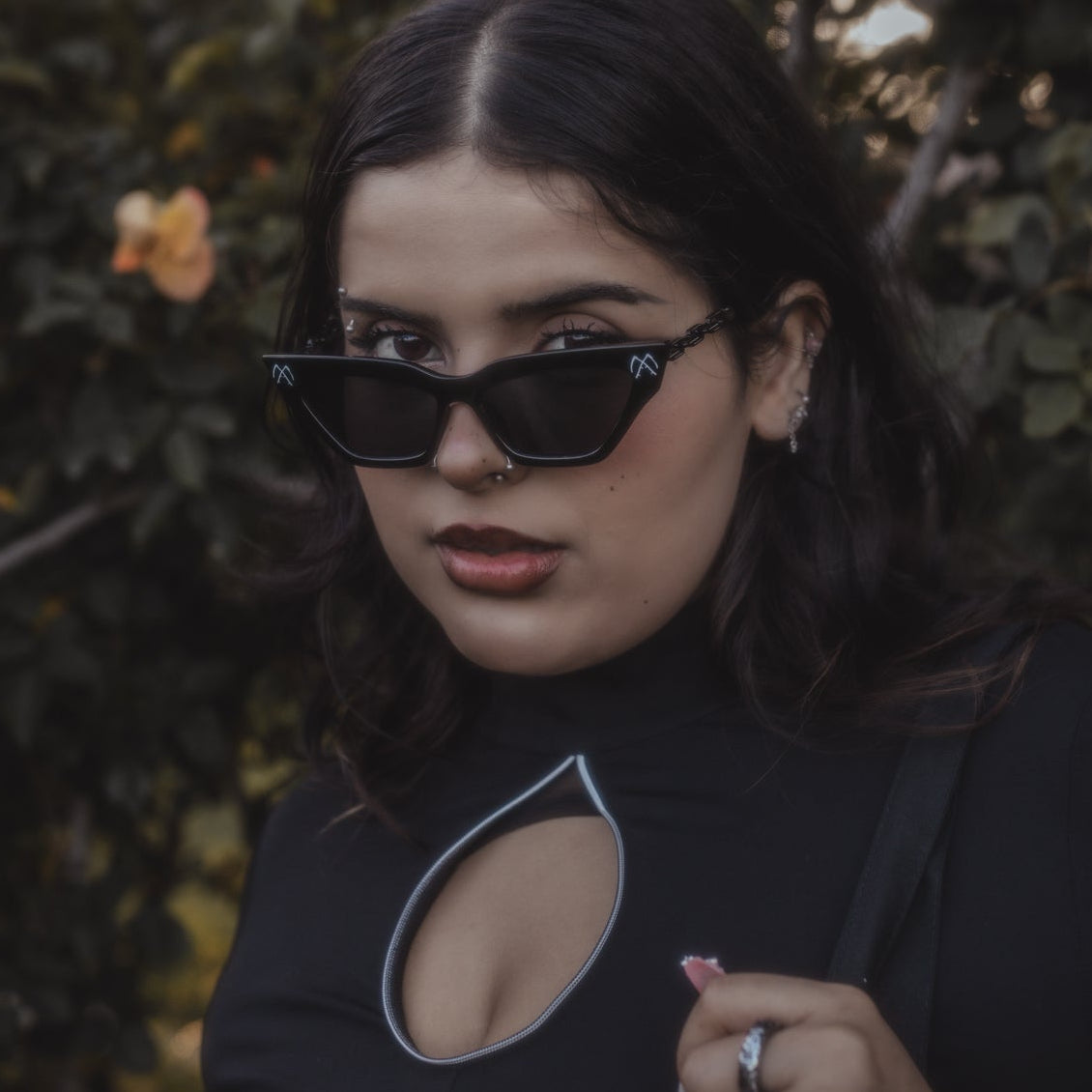 Woman wearing sunglasses and a black outfit with cut-out design, standing against a dark background.