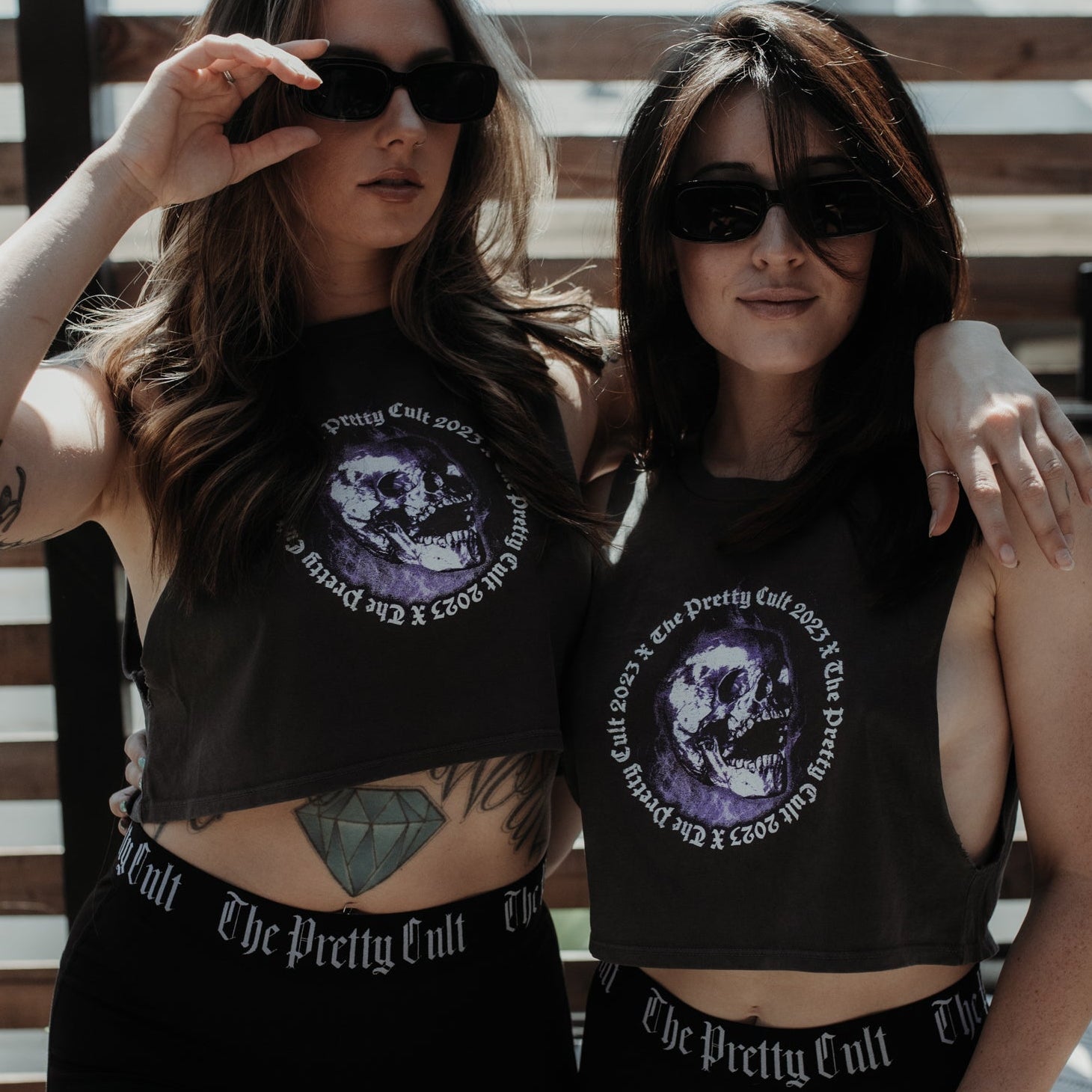 Two women wearing matching black crop tops and shorts with a graphic design, standing against a wooden background.