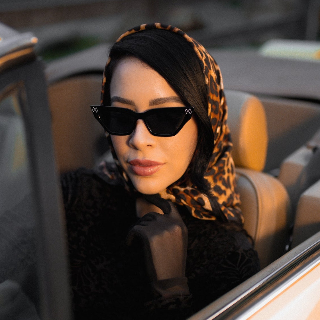 Woman wearing sunglasses and a leopard print headscarf sitting in a car.