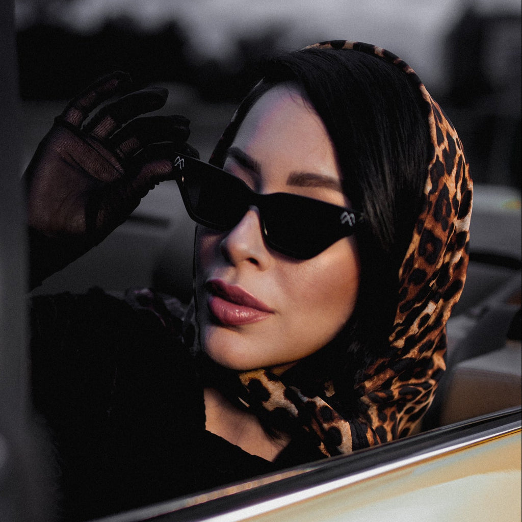 Woman wearing sunglasses and a leopard print headscarf sitting in a car.