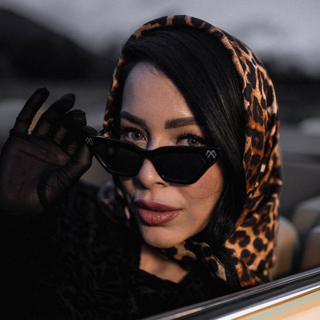 Woman wearing sunglasses and a leopard print headscarf, sitting in a car.