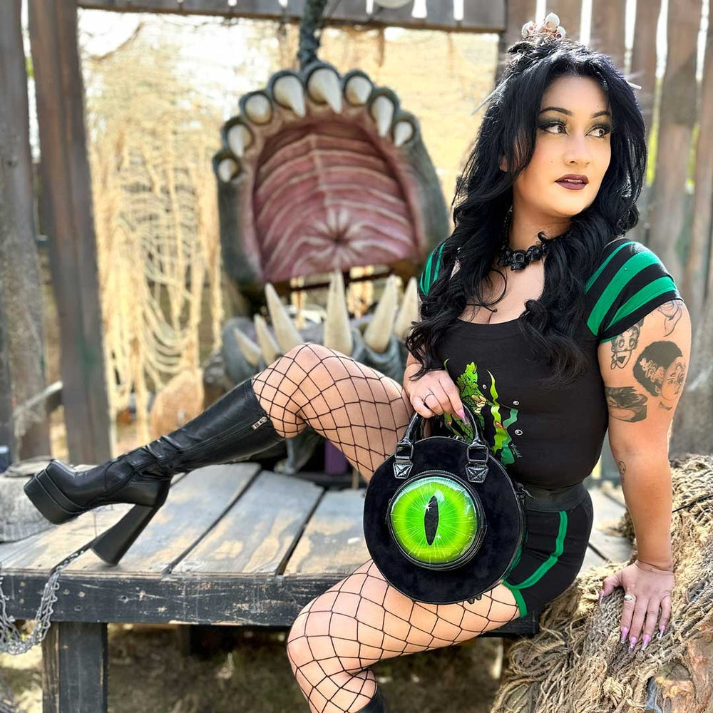 Woman in a black and green outfit with a large eye-shaped bag, sitting outdoors.