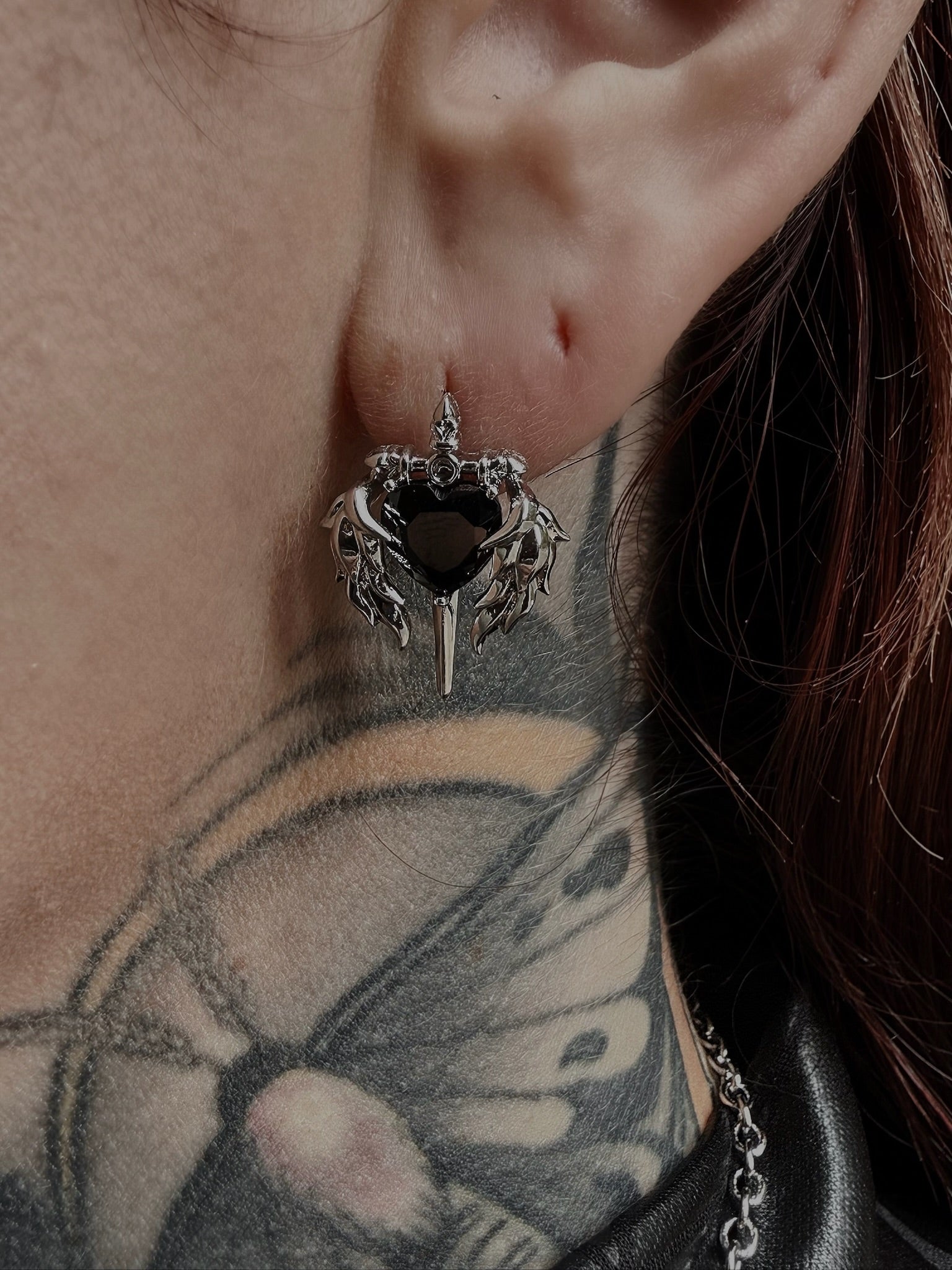 Close-up of an ear with a black heart-shaped earring featuring wings.