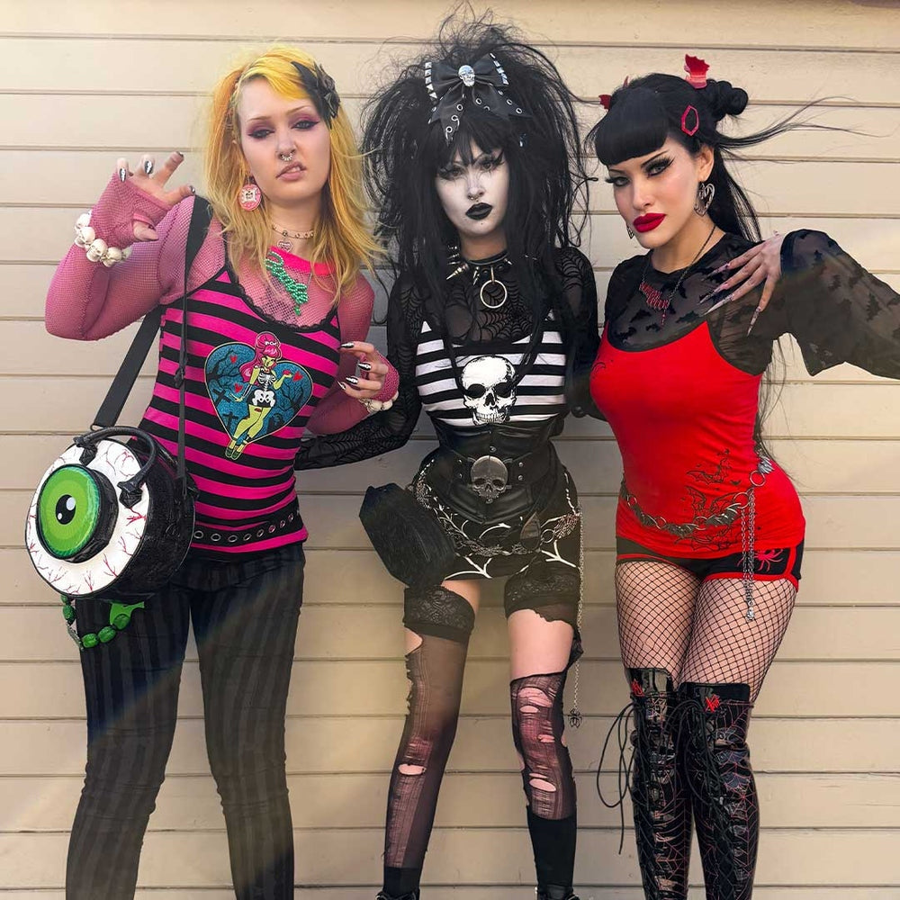 Three women in gothic and punk-inspired outfits posing against a wooden wall.