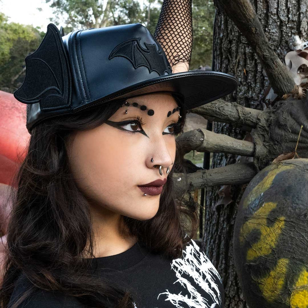 A woman wearing a black faux-leather witch-style hat with bat wings on the brim and a horn, styled with a fishnet headband, in an outdoor Halloween setting.
