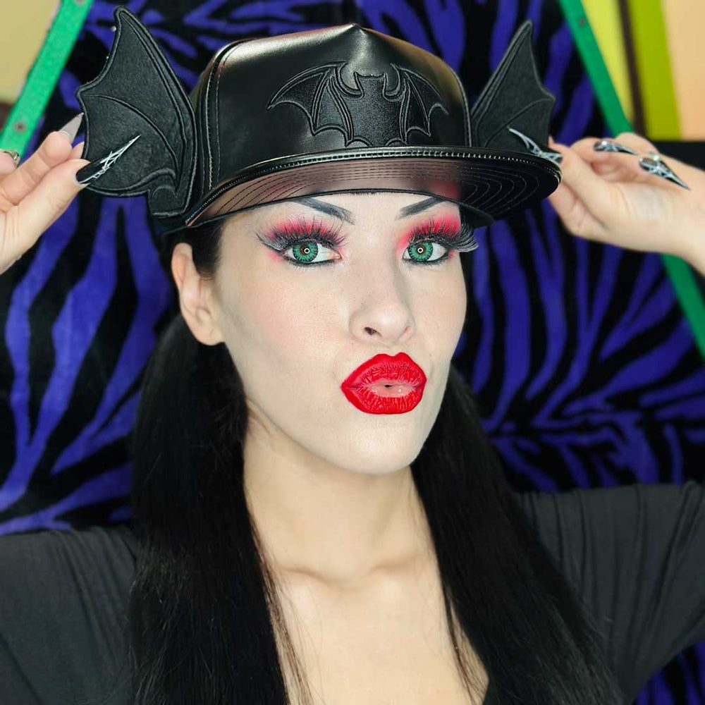 A woman wearing a black faux-leather gothic-style cap with bat wings on the sides and a bat emblem on the front, posing against a purple and black zebra-striped background.