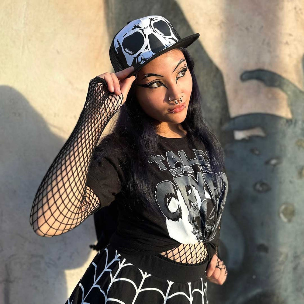 A woman posing in a gothic streetwear outfit, featuring a black and white skull print baseball cap, a black graphic t-shirt, fishnet arm sleeves, and a black and white spiderweb-patterned skirt, set against a textured concrete wall with dramatic shadows.
