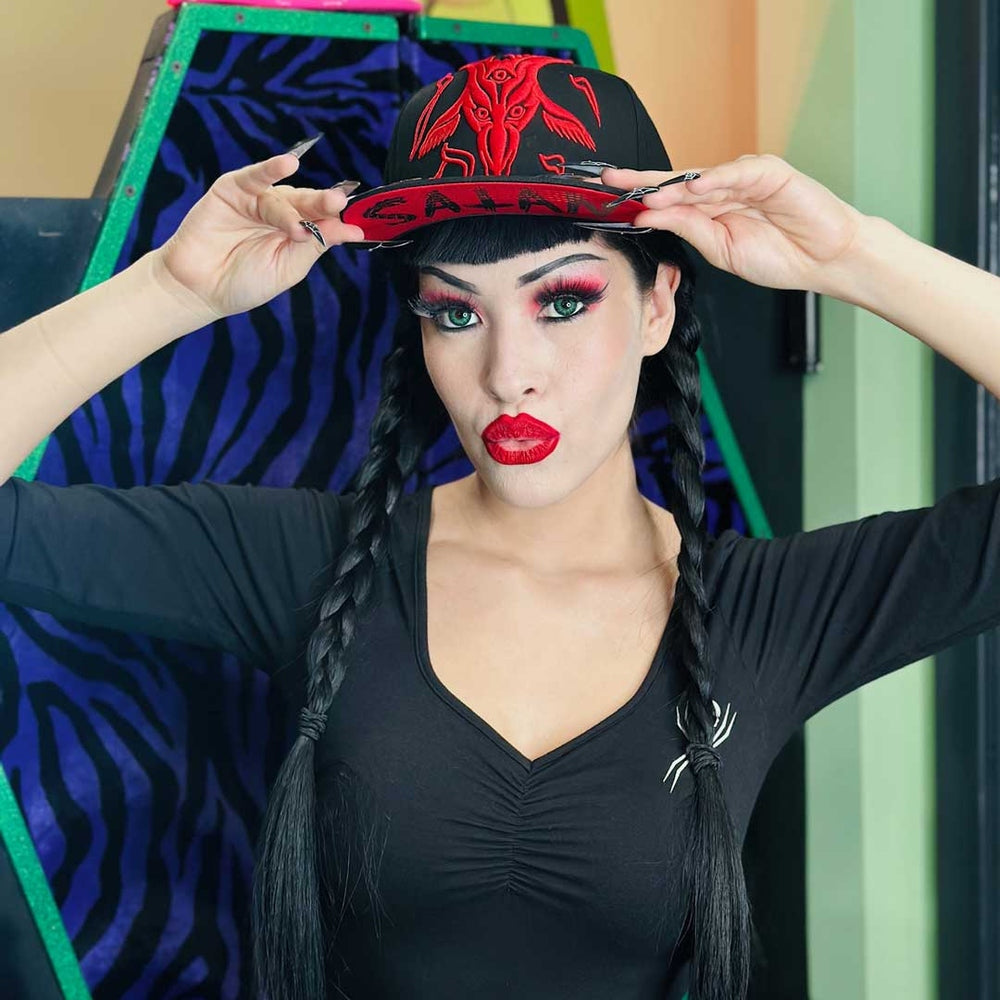 A woman with gothic makeup, long black braids, and a black cap with red embroidery and the word 'SATAN' on the front. She is wearing a black V-neck top with a white spider graphic, holding the brim of the hat.