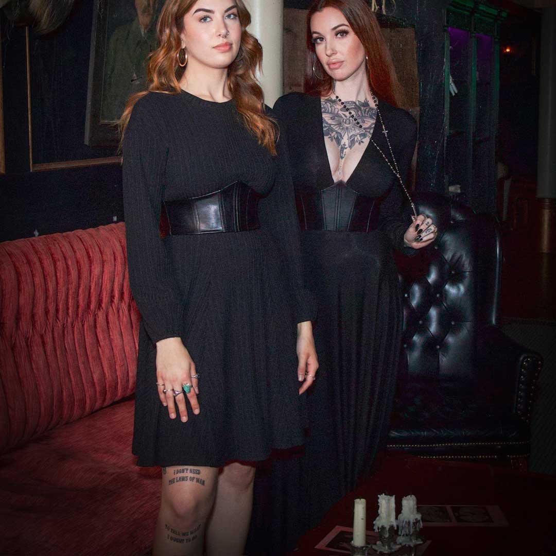 Two women in black dresses standing in a dimly lit room with red couches and candles.