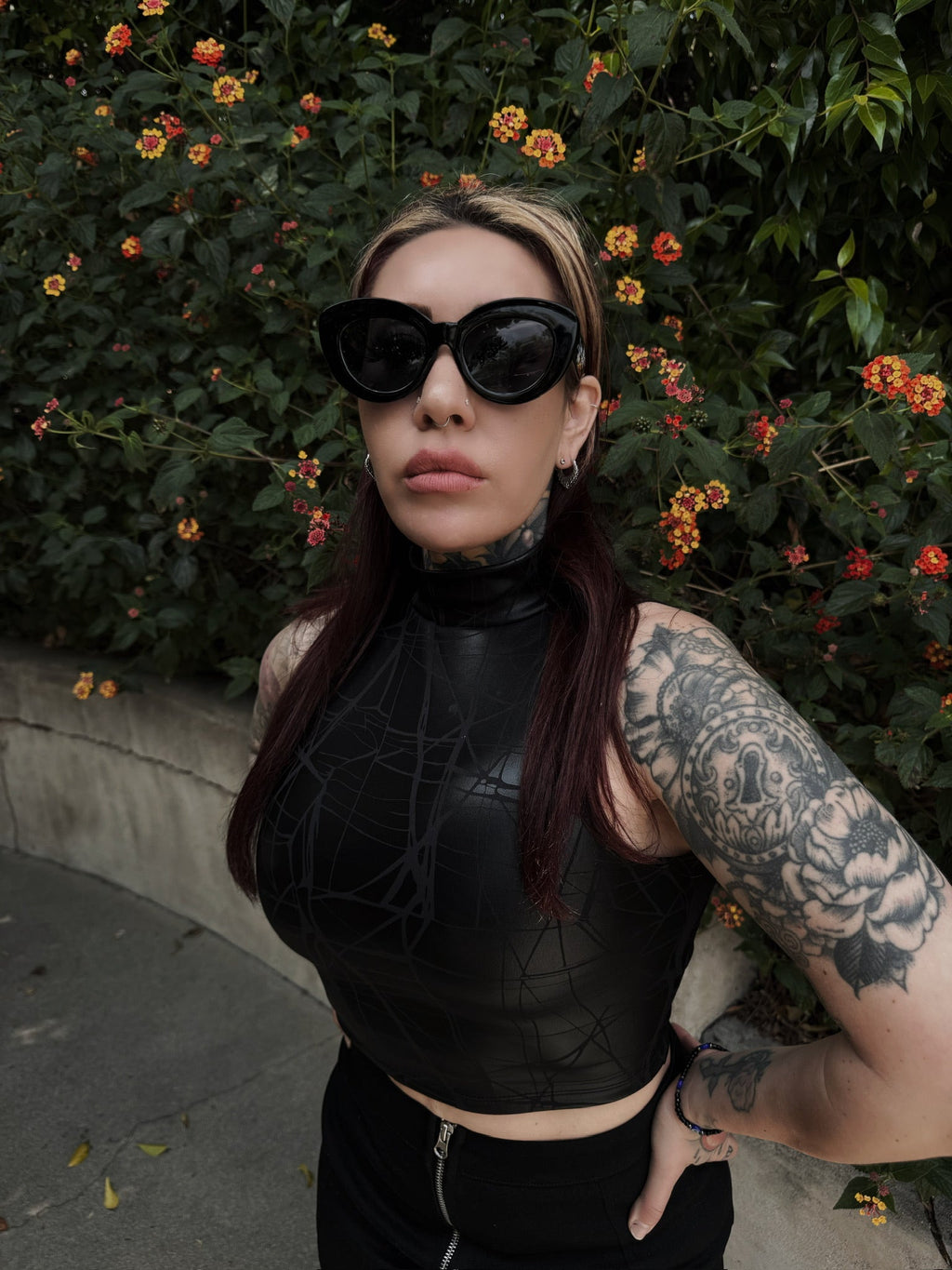 Person wearing a black textured top with tattoos, standing in front of green foliage with flowers.