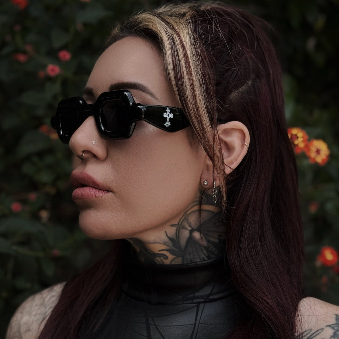 Person with tattoos wearing sunglasses and a black outfit against a natural background