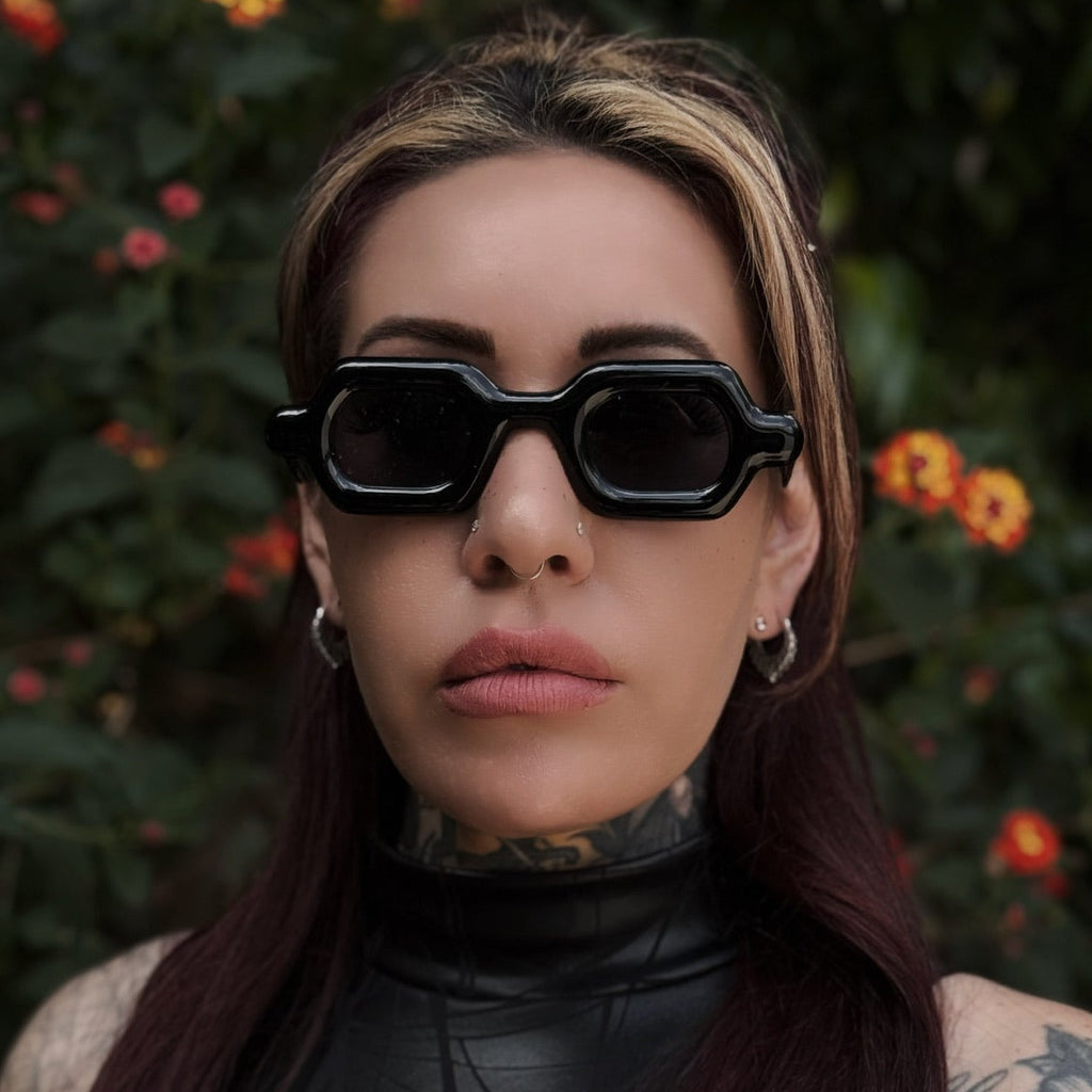Person wearing black sunglasses and a black outfit with tattoos, standing against a natural background with flowers.