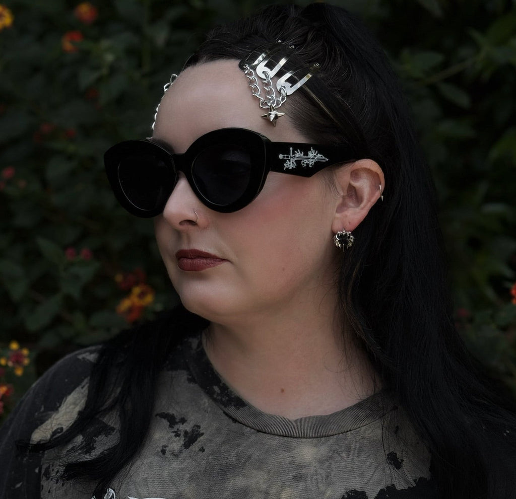 Person wearing dark sunglasses and a black outfit with intricate designs, standing against a blurred natural background.