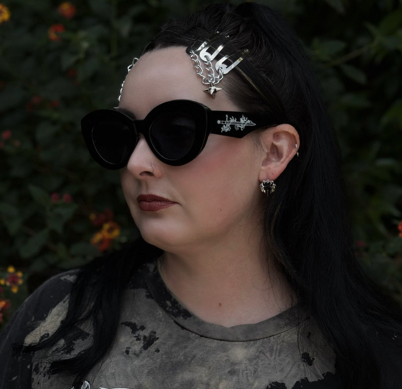 Person wearing dark sunglasses and a black outfit with intricate designs, standing against a blurred natural background.