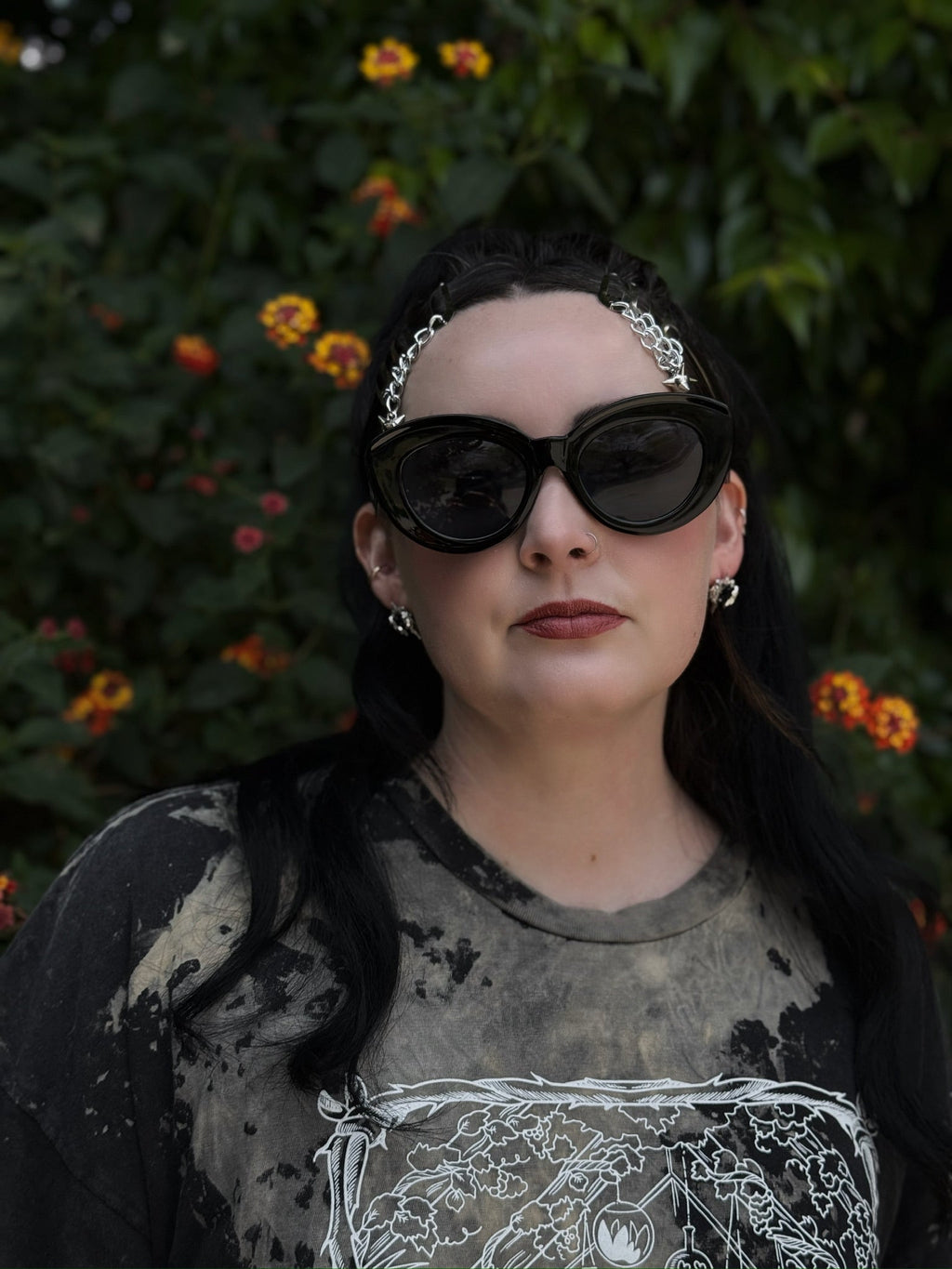 Person wearing sunglasses and a patterned shirt with a dark background and flowers.