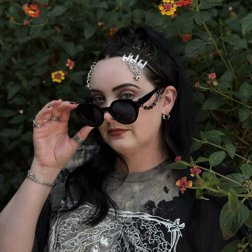 Person with dark hair and sunglasses standing among green foliage and flowers