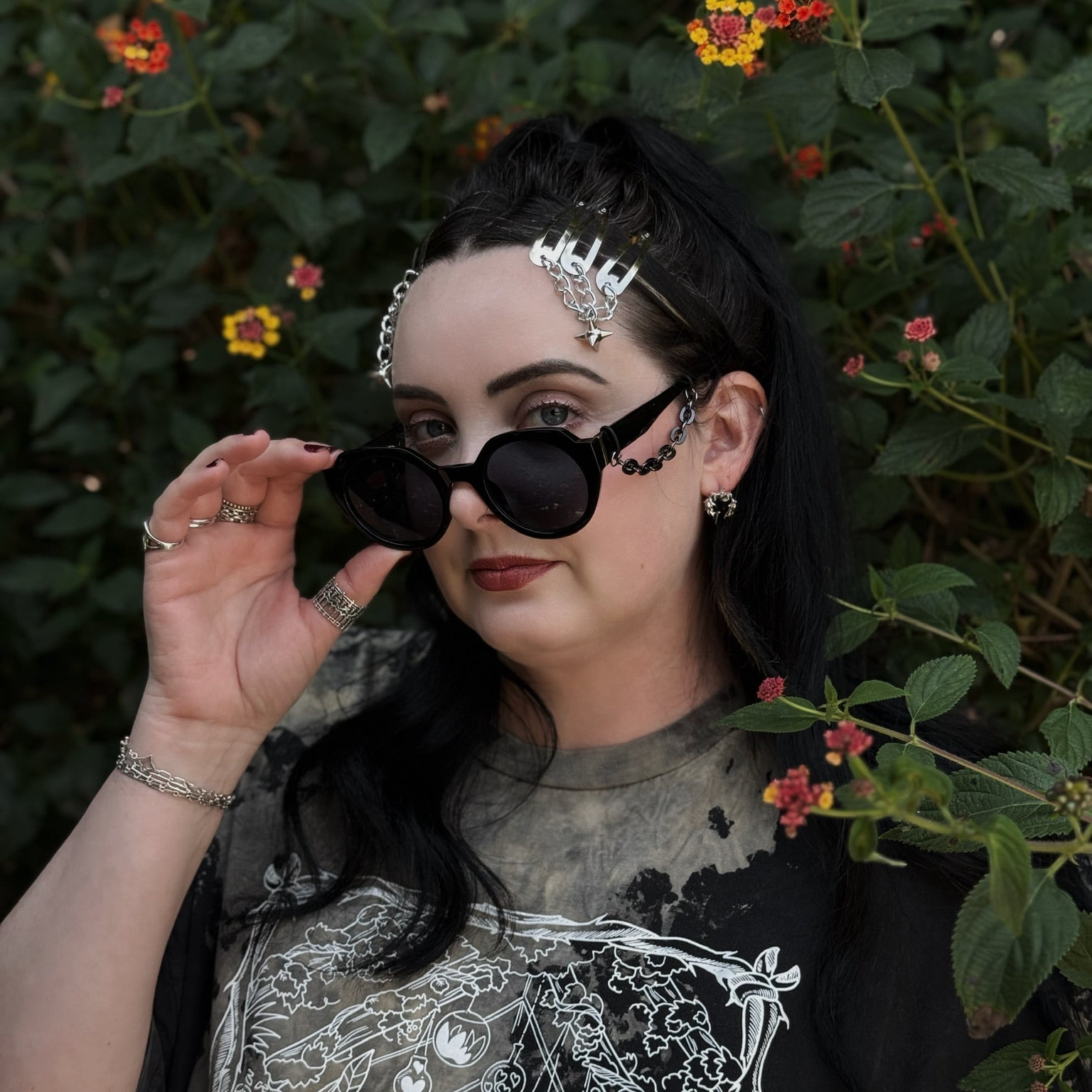 Person with dark hair and sunglasses standing among green foliage and flowers