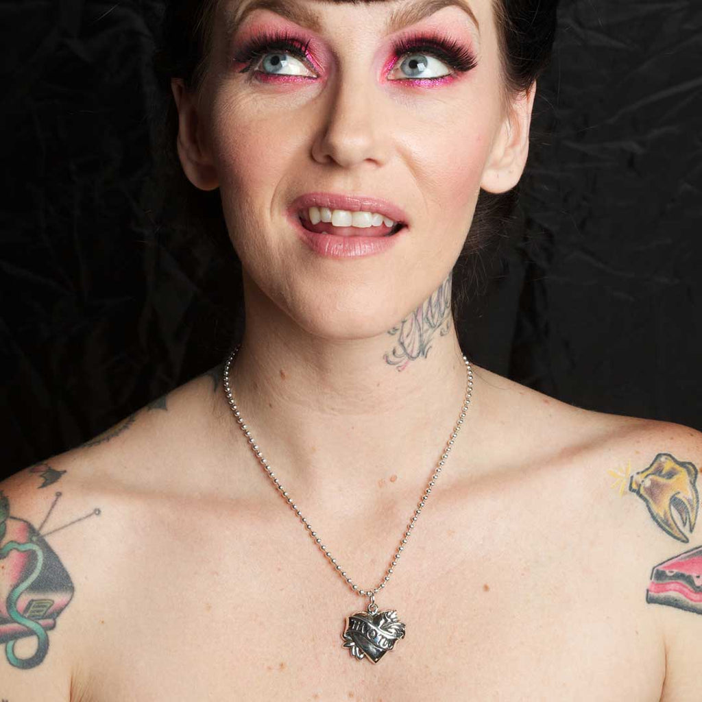 Woman with tattoos and pink makeup wearing a necklace against a dark background