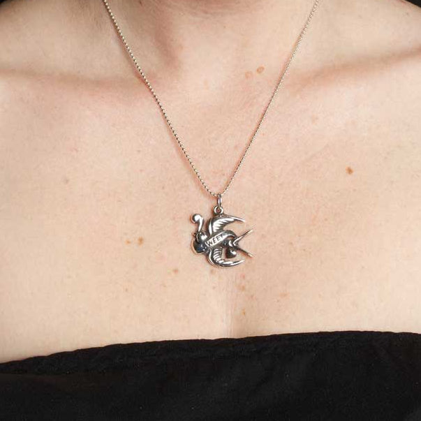 Person wearing a necklace with a unique pendant, surrounded by tattoos on arms and shoulders.