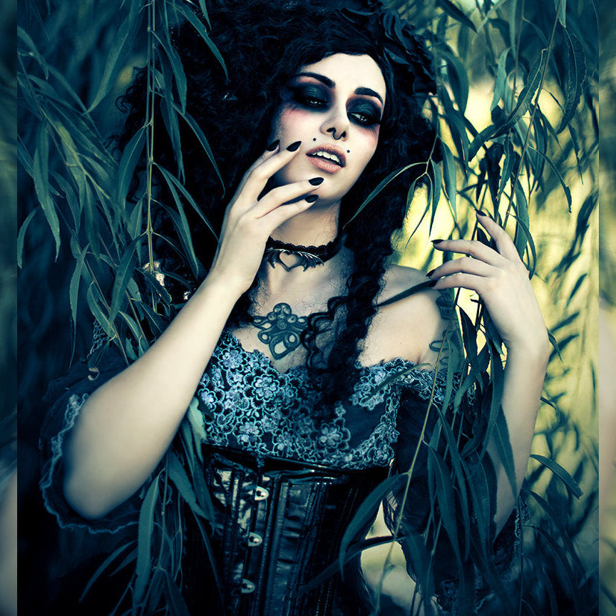 Woman in a dark, ornate dress amidst green foliage with a mystical atmosphere.