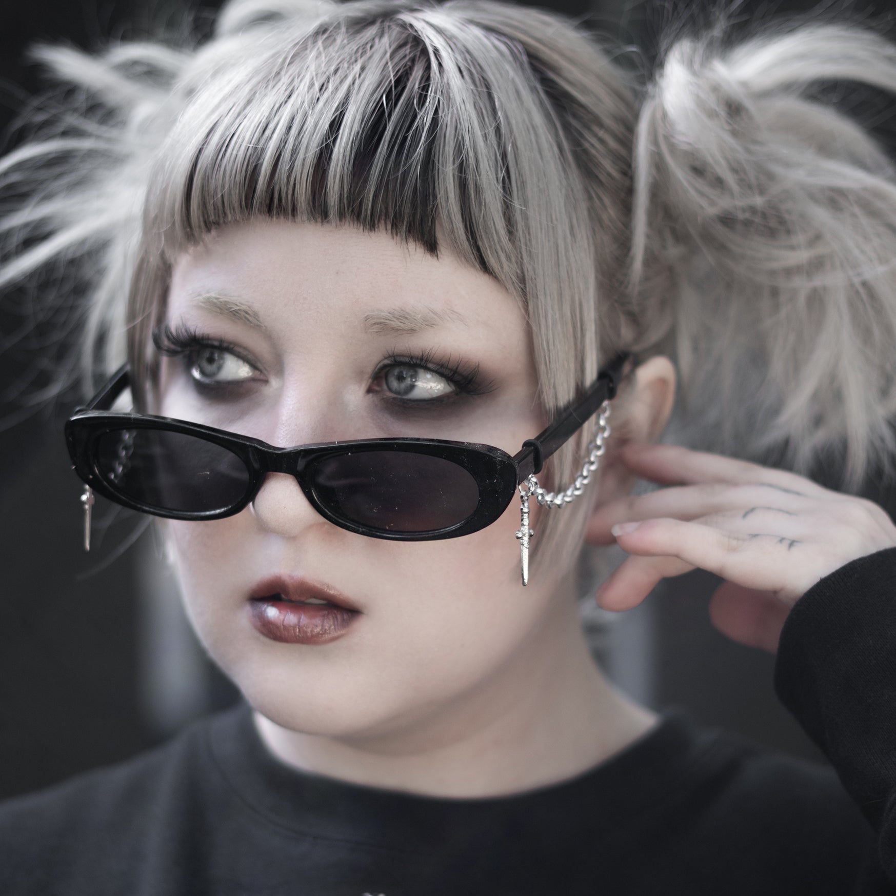 Person with styled hair and sunglasses, wearing a black sweater with a white emblem.