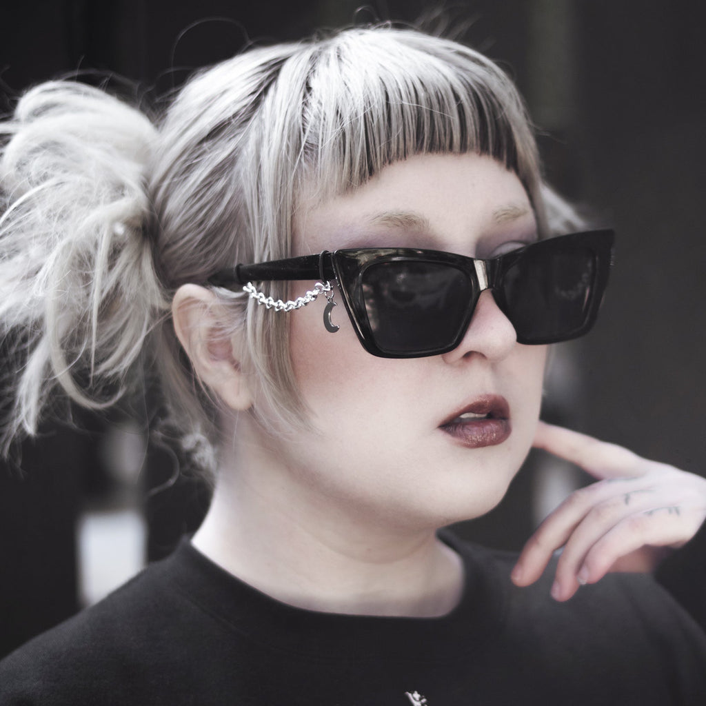 Person wearing black sunglasses with a decorative chain, black shirt with emblem, and blonde hair styled in pigtails.