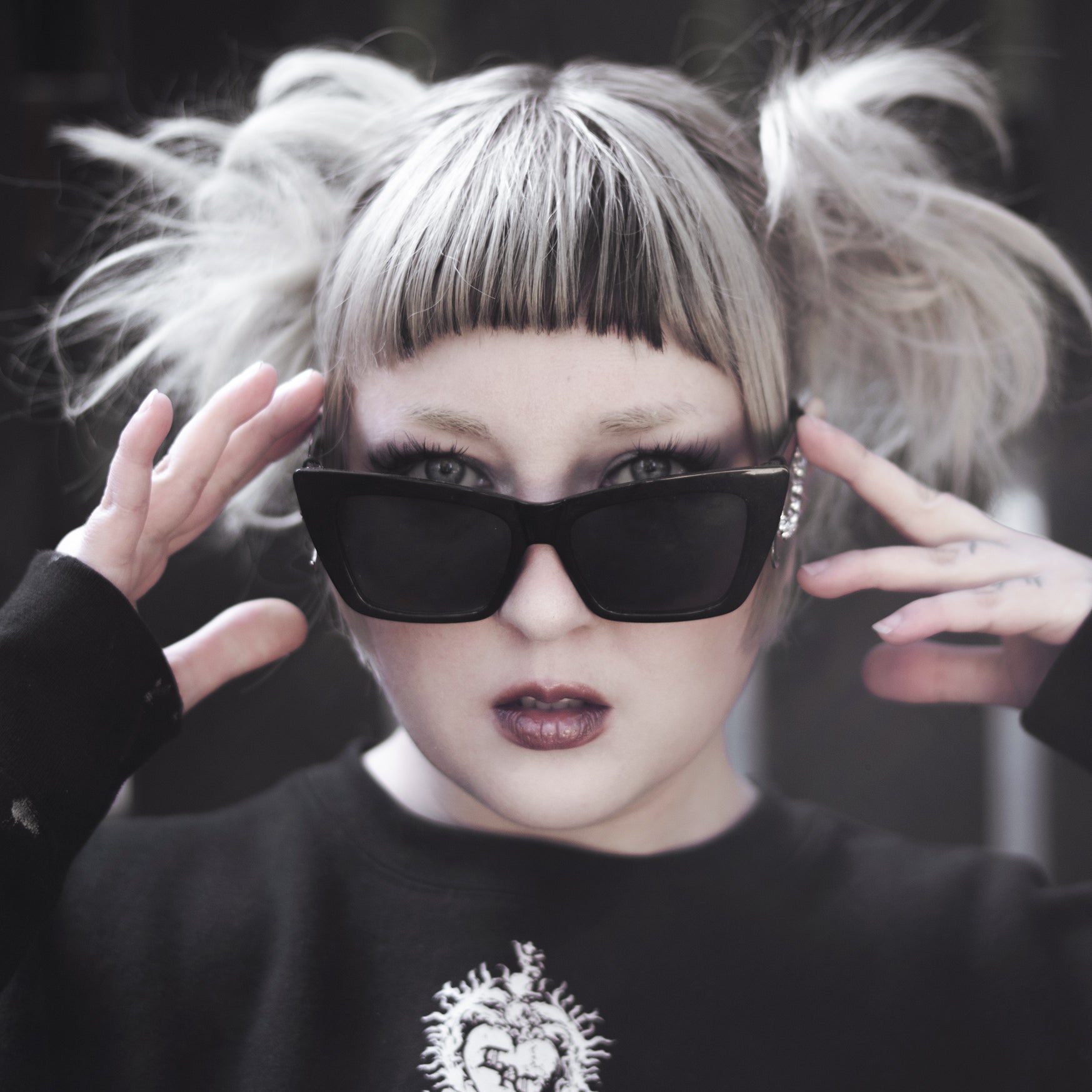 Person with styled hair and black sunglasses wearing a black sweatshirt with a heart design.