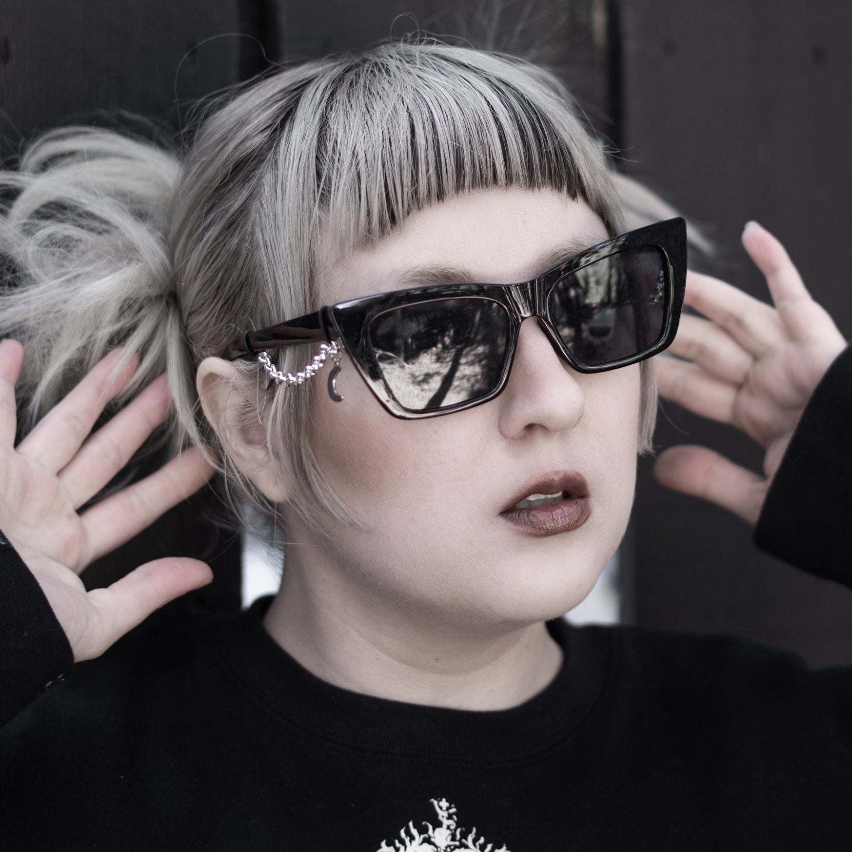 Person wearing large sunglasses with a black sweater featuring a white emblem.