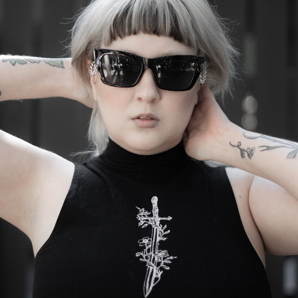Person wearing a black sleeveless top with a white design, sunglasses, and tattoos.