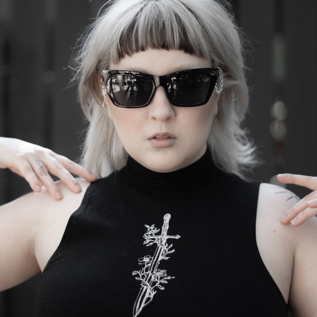 Person wearing a black sleeveless top with a white design, sunglasses, and a blurred background.