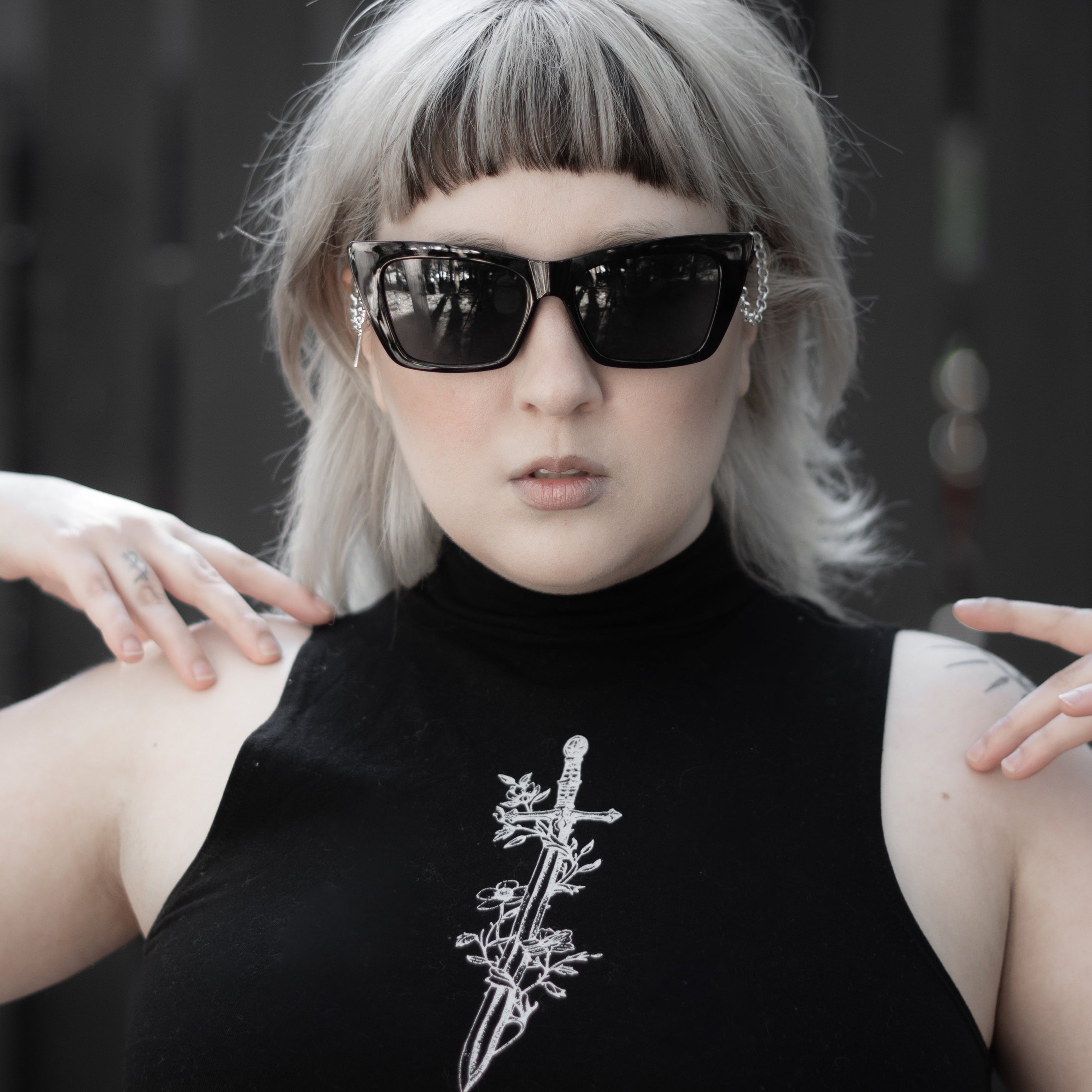 Person wearing a black sleeveless top with a white design, sunglasses, and a blurred background.