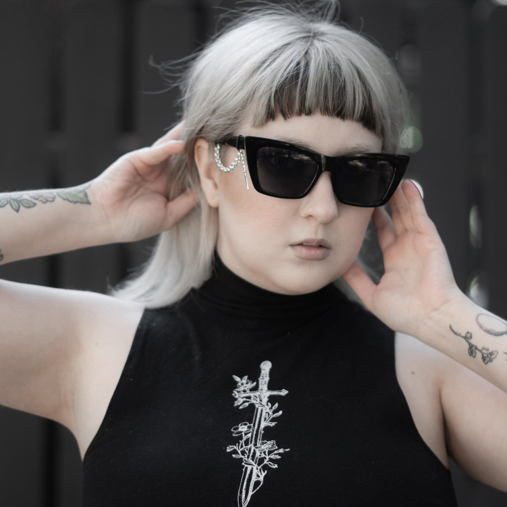 Person wearing a black sleeveless top with a white design, sunglasses, and holding their hair back.