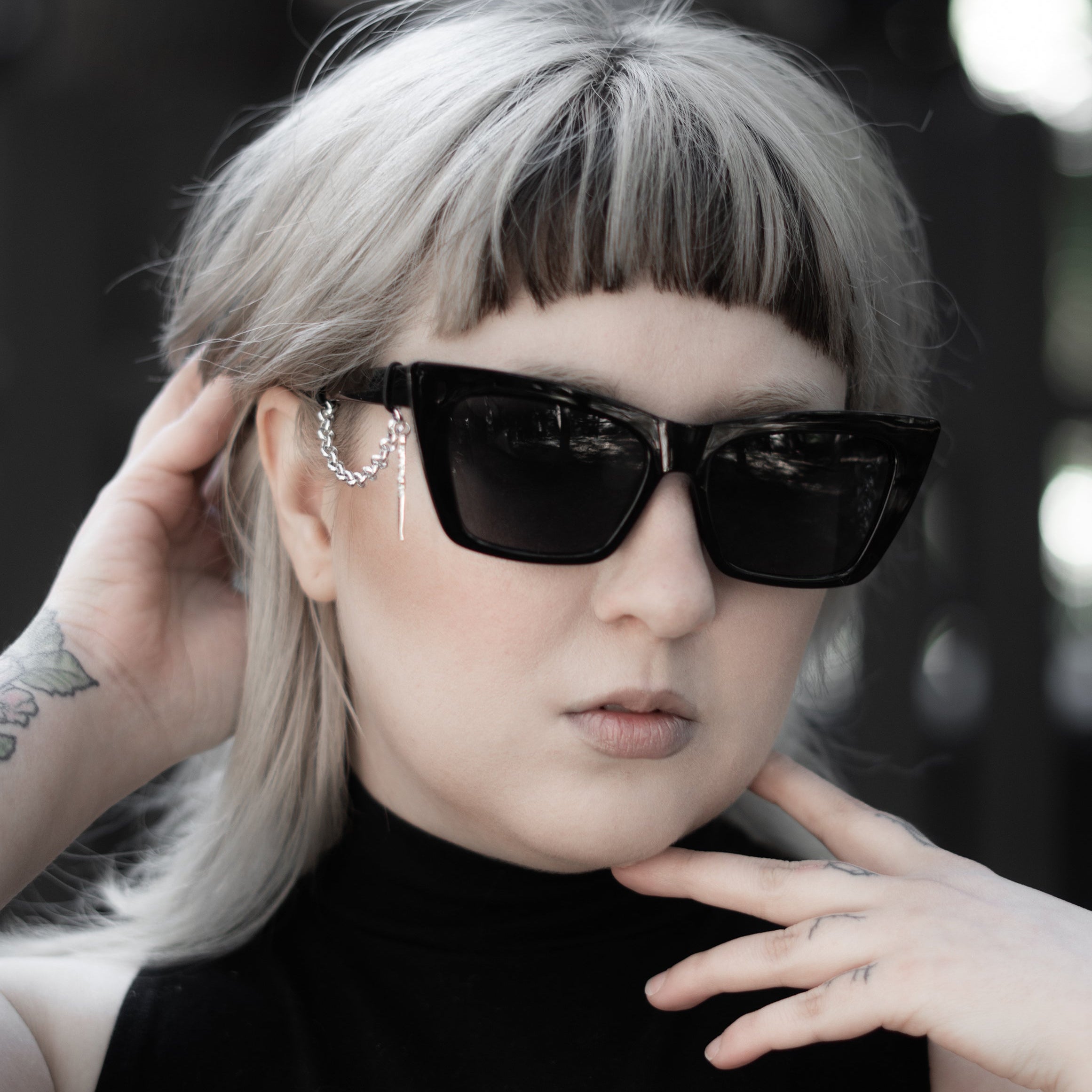 Person with short blonde hair wearing black sunglasses and a black sleeveless top with white design.