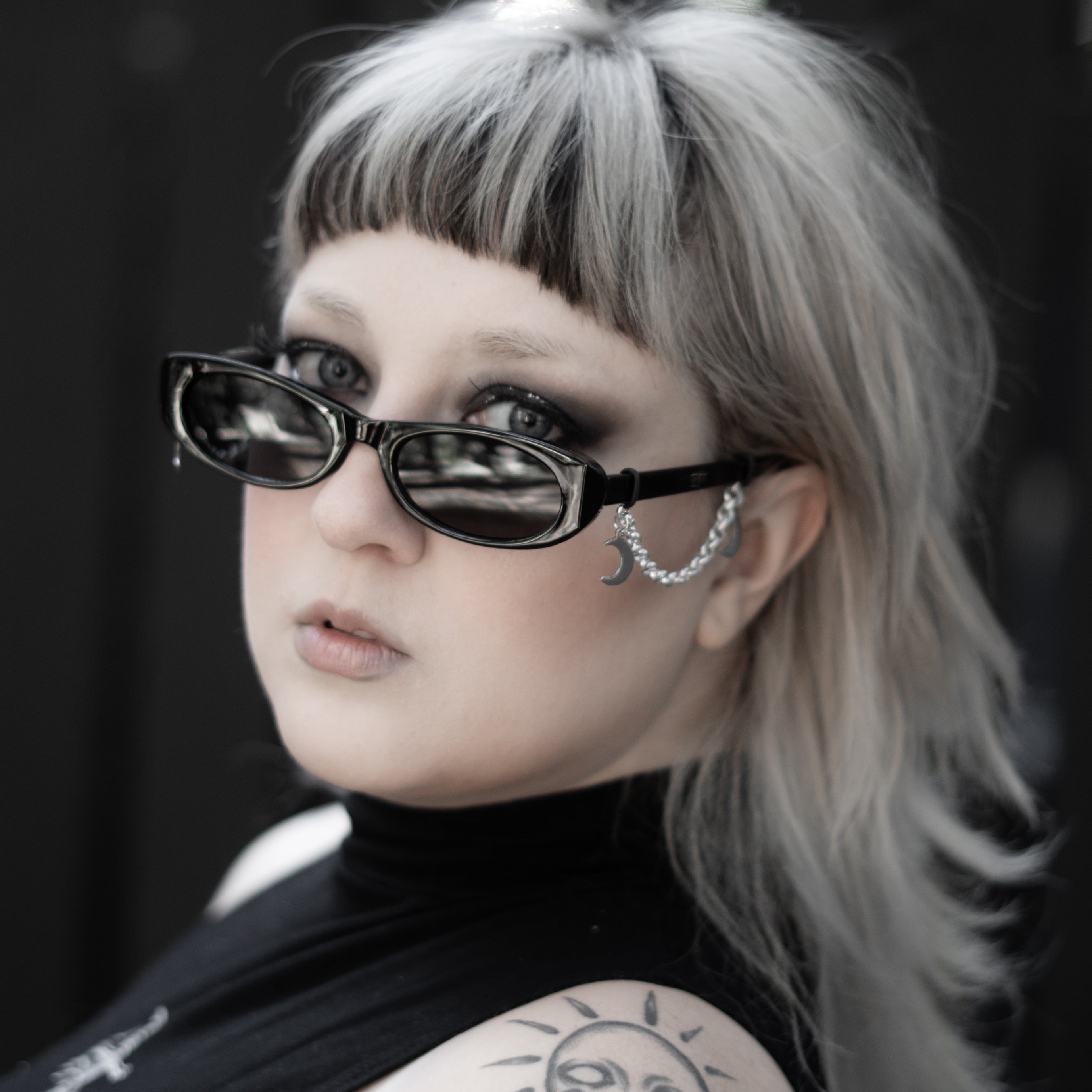 Person with sunglasses and a sun tattoo on shoulder against a dark background