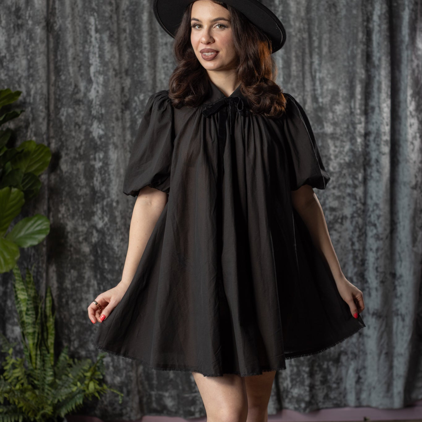 Woman wearing a black dress and hat against a textured wall.