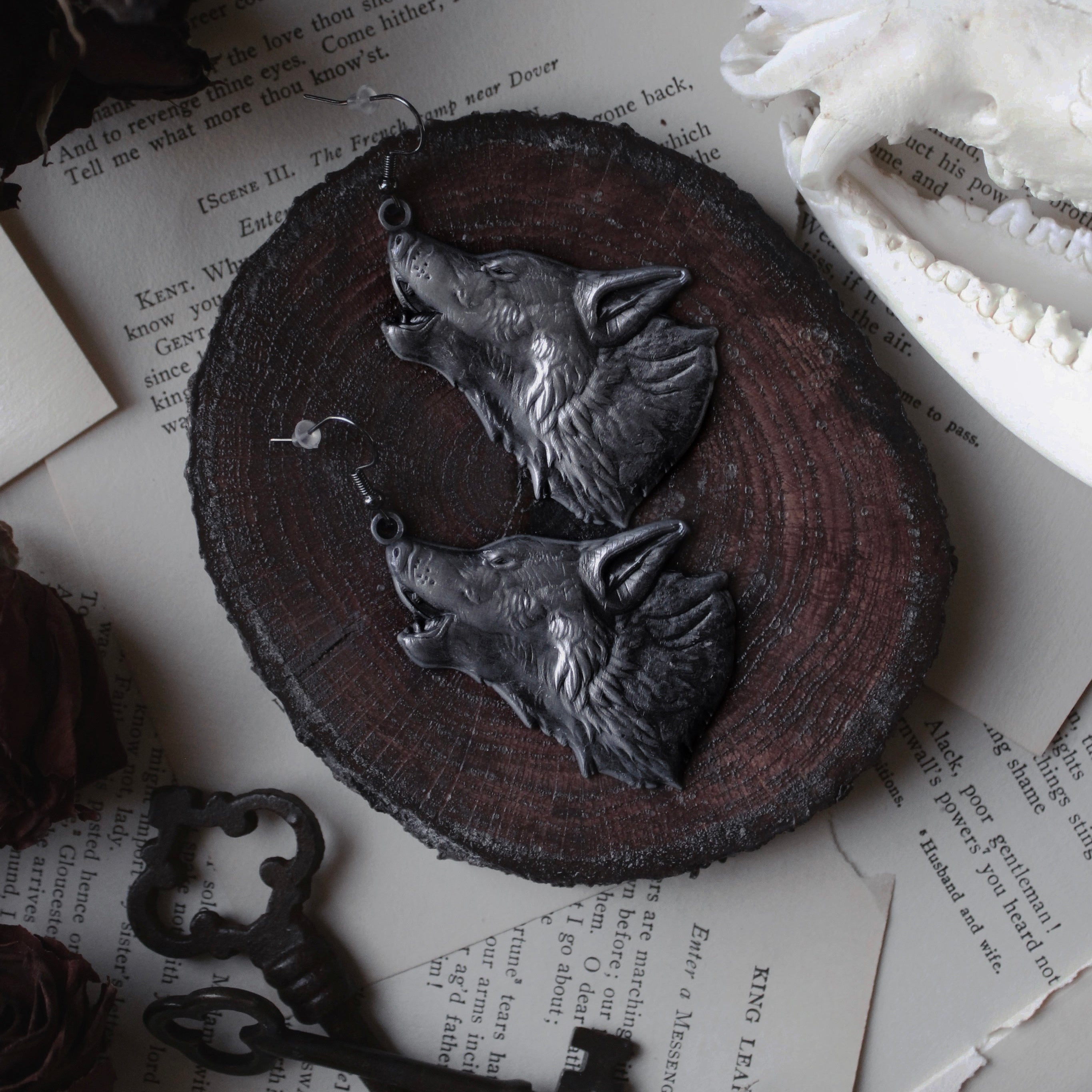 Wolf heads earrings on a wooden base, surrounded by dried flowers and text.