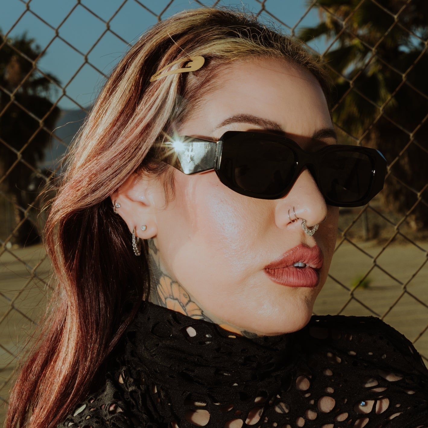 Woman wearing sunglasses and a black lace top with a chain-link fence and palm trees in the background.