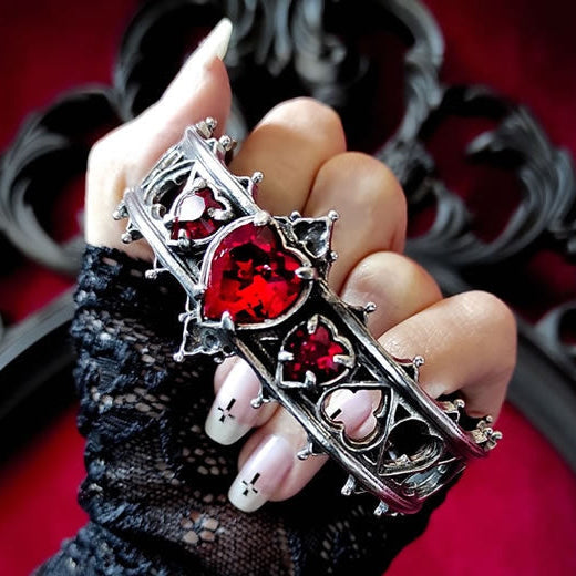 A hand wearing a black lace glove holds a gothic-style silver cuff bracelet adorned with red heart-shaped crystals and small white heart details, set against a dark ornate frame and red background.