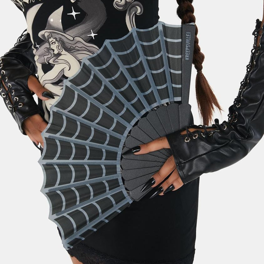 A gothic-style model wearing a black lace dress and matching fingerless gloves, holding a gray folding fan with a spiderweb pattern. The dress features a graphic of a witch and the word 'Halloween'.