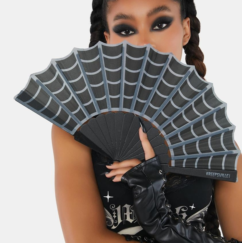 A model holds a black folding fan with a gray spiderweb pattern, wearing a black graphic top and black lace-up arm warmers against a light gray background.