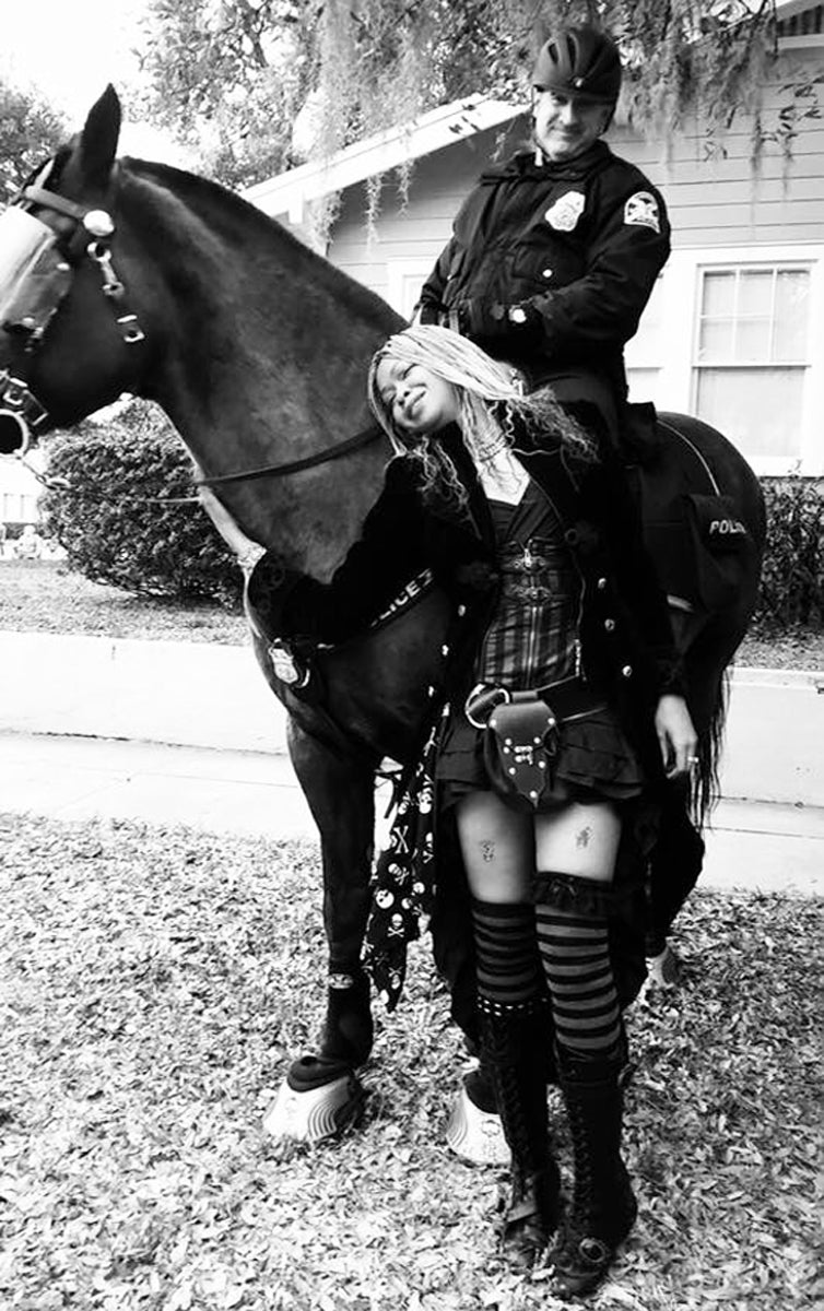 Person in a plaid skirt and striped stockings standing next to a horse, with another person on horseback in the background.