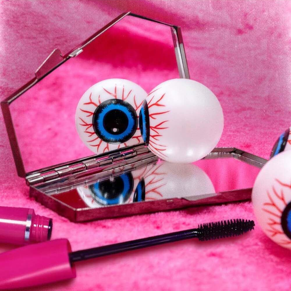 Glabellum eyelash extensions with realistic white, red, and blue eyeballs attached to black lash bands, shown with a pink mascara tube and wand on a pink textured background.