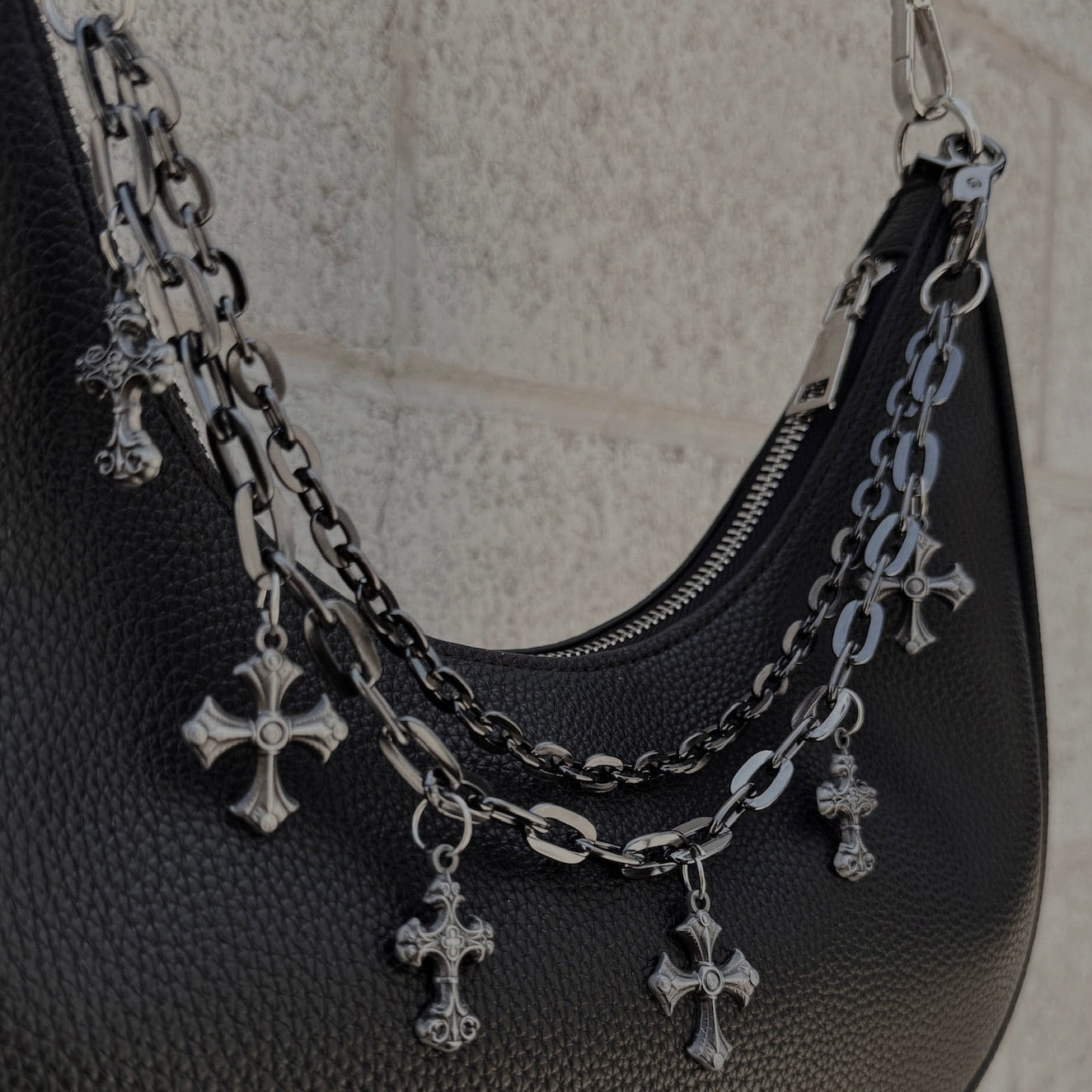 Black handbag with a silver chain and cross charm against a light stone wall.