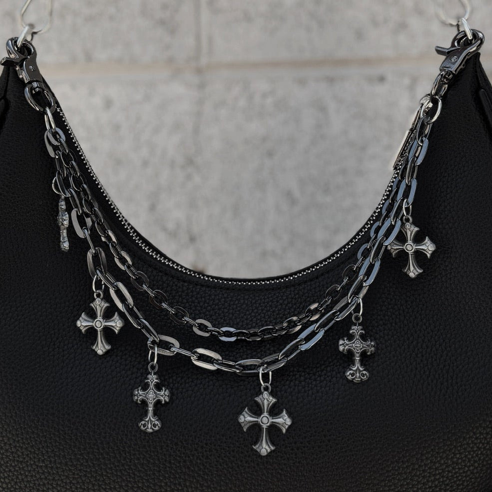 Black handbag with silver chain and cross charm held by a person against a gray tiled wall.